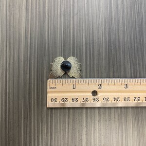 Może przedstawiać: Złota przypinka w kształcie anielskich skrzydeł, z czarnym tyłem. Przypinka ma szerokość około 2,5 cm. Przypinka leży na szarej drewnianej powierzchni obok linijki.