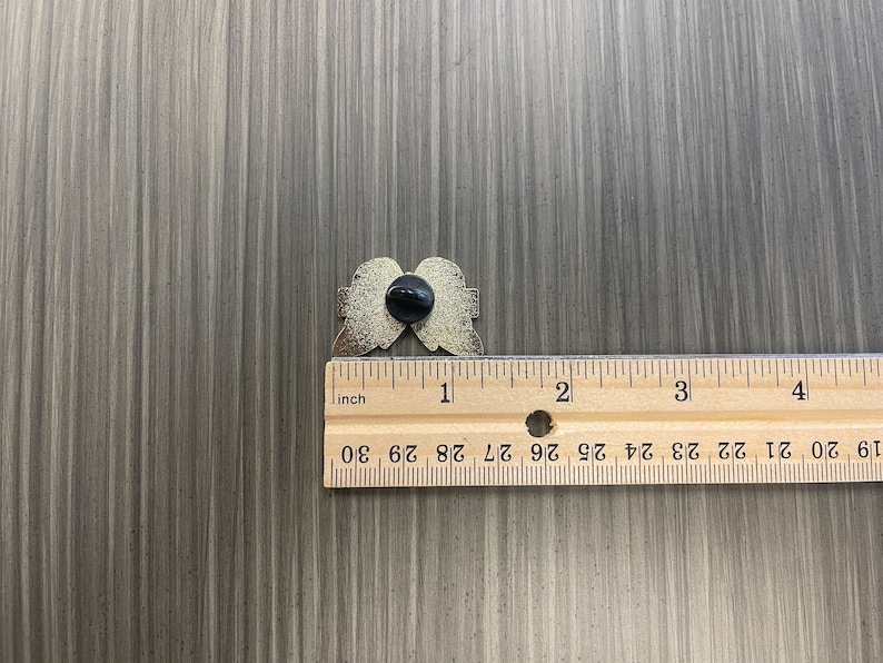 Może przedstawiać: Złota przypinka w kształcie anielskich skrzydeł, z czarnym tyłem. Przypinka ma szerokość około 2,5 cm. Przypinka leży na szarej drewnianej powierzchni obok linijki.