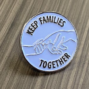 May include: White enamel pin with a silver border. The pin features two hands holding each other and the text "KEEP FAMILIES TOGETHER".