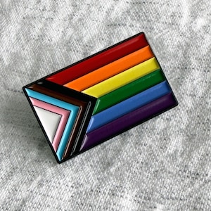 May include: A rectangular enamel pin featuring the Progress Pride flag design. The pin displays horizontal stripes in red, orange, yellow, green, blue, and purple, with a black and brown chevron on the left side containing white, pink, and light blue stripes.