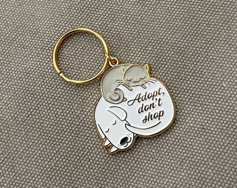 May include: Gold key chain with a white dog and a gray cat. The dog is holding the cat and the text "Adopt, don't shop" is on the dog's body.