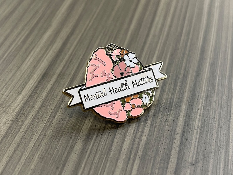 May include: A gold-toned pin featuring a pink brain design with floral accents. A white banner across the brain reads "Mental Health Matters" in black lettering. The pin is set against a gray wooden surface.