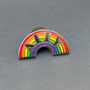 May include: A rainbow-colored enamel pin with the word "ALLY" in black letters. The pin is in the shape of a rainbow with red, orange, yellow, green, blue, and purple stripes. The pin has a silver border.