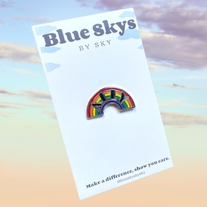 May include: A rainbow-colored enamel pin with the word "ALLY" in black letters, set against a rainbow arch. The pin is on a white card with the text "Blue Skys by Sky" and "Make a difference, show you care."