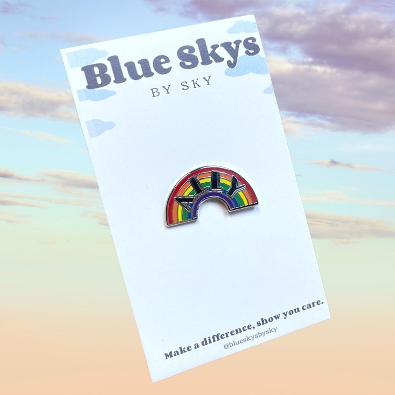 May include: A rainbow-colored enamel pin with the word "ALLY" in black letters, set against a rainbow arch. The pin is on a white card with the text "Blue Skys by Sky" and "Make a difference, show you care."