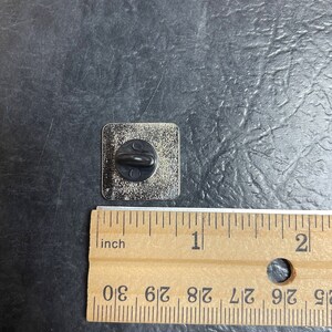May include: A square metal pin with a black post on the back. The pin is silver with a slightly textured surface. A wooden ruler is next to the pin, showing measurements in inches.