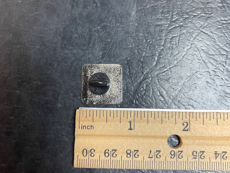 May include: A square metal pin with a black post on the back. The pin is silver with a slightly textured surface. A wooden ruler is next to the pin, showing measurements in inches.