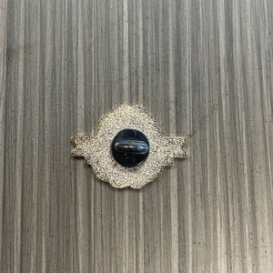 May include: Back of a gold-colored pin with a black pin back. The pin has a decorative, ornate design with a textured surface. The pin is on a gray wooden surface.