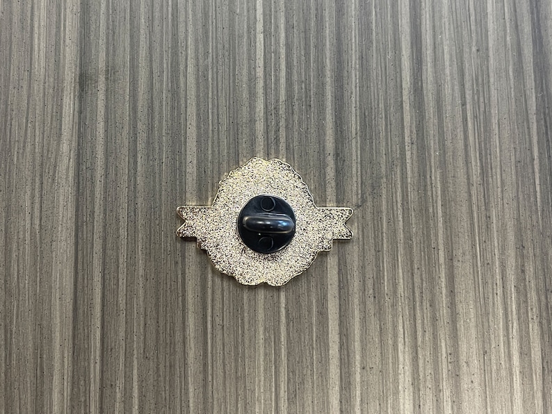 May include: Back of a gold-colored pin with a black pin back. The pin has a decorative, ornate design with a textured surface. The pin is on a gray wooden surface.
