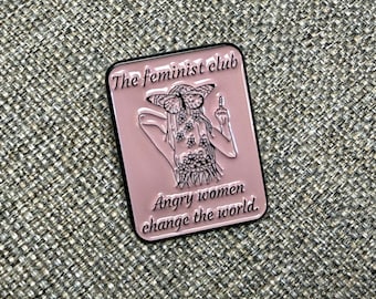 May include: A pink enamel pin with a black border. The pin features an illustration of a figure with butterfly wings and floral details, giving the middle finger. Text on the pin reads "The feminist club" and "Angry women change the world."