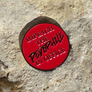 May include: A red, circular button with black text that reads "IMPEACH THE PEVOPHILE IN OFFICE." The button has a black border and is set against a textured, light-colored stone surface.