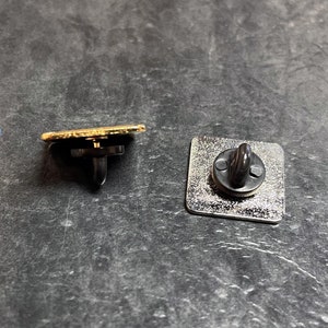May include: Two metal pin backs. One is a gold-edged rectangle with a black post and clutch. The other is a silver square with a black post and clutch. Both are on a dark gray surface.
