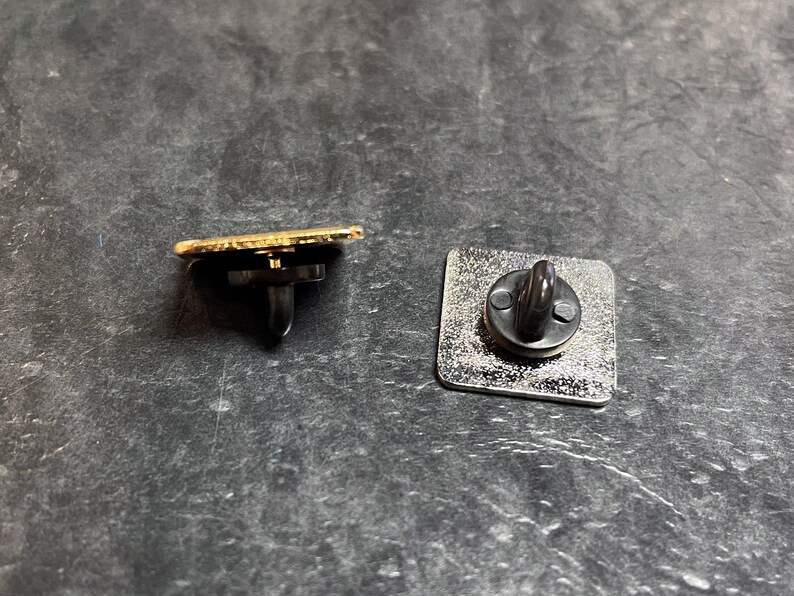 May include: Two metal pin backs. One is a gold-edged rectangle with a black post and clutch. The other is a silver square with a black post and clutch. Both are on a dark gray surface.