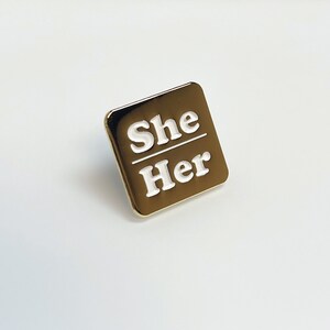 May include: A gold-colored square pin with the words "She" over "Her" in white lettering. The pin has a shiny, metallic finish and a simple, modern design. The pin is likely a pronoun badge.