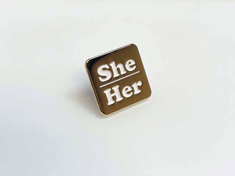 May include: A gold-colored square pin with the words "She" over "Her" in white lettering. The pin has a shiny, metallic finish and a simple, modern design. The pin is likely a pronoun badge.