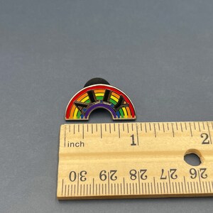 May include: A rainbow-colored enamel pin with the word "ALLY" in black letters. The pin is in the shape of a rainbow and measures approximately 1 inch in length. The pin is on a gray surface.
