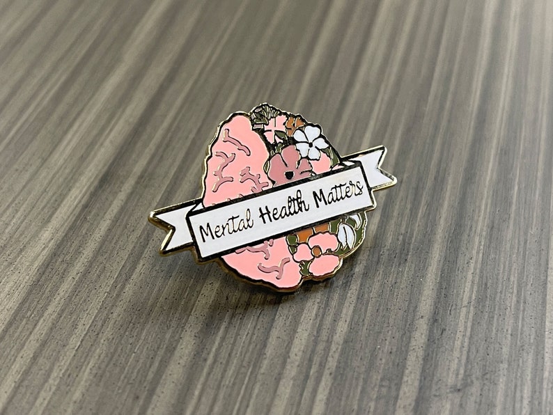 May include: A gold-toned pin featuring a pink brain design with floral accents. A white banner across the brain reads "Mental Health Matters" in black lettering. The pin is set against a gray wooden surface.