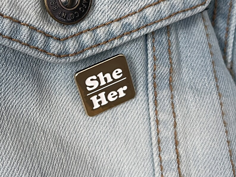 May include: A square, bronze-colored enamel pin with the words "She Her" in white, block letters. The pin is affixed to the pocket of a light blue denim jacket. The jacket has a metal button.