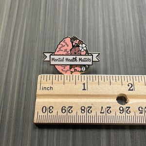 May include: A pin featuring a stylized brain design with floral accents in pink, brown, and white. The pin includes a banner that reads "Mental Health Matters". The pin is approximately 1 inch in length.
