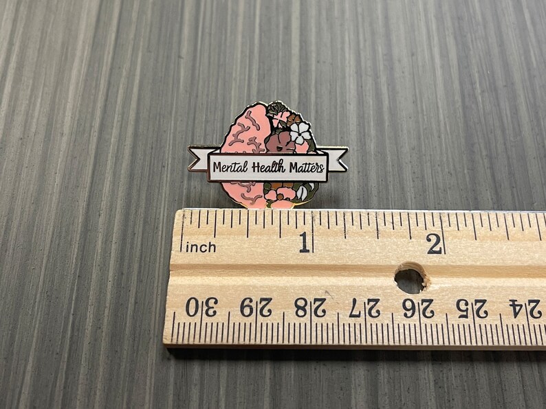 May include: A pin featuring a stylized brain design with floral accents in pink, brown, and white. The pin includes a banner that reads "Mental Health Matters". The pin is approximately 1 inch in length.