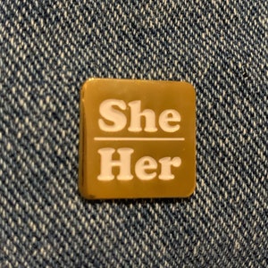 May include: A square, gold-colored pin with the words "She" over "Her" in white, sans-serif font. The pin is set against a blue denim background.