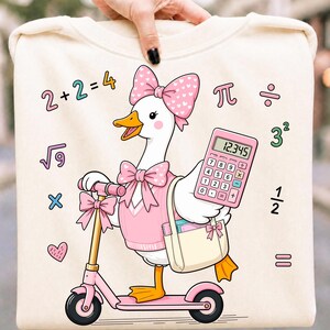 May include: A cream-coloured sweatshirt featuring a cartoon goose wearing a pink sweater and bow, riding a pink scooter. The goose holds a pink calculator and a tote bag. Mathematical symbols and equations like 2+2=4 are printed around the goose.