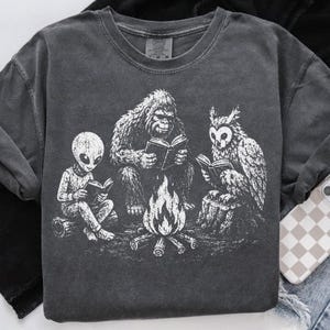 May include: A charcoal gray crew neck t-shirt featuring a white graphic print of an alien, a Bigfoot, and an owl sitting around a campfire while reading books. The shirt has a vintage-style washed finish and a standard short-sleeve design.