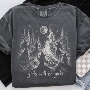Può includere: Una t-shirt girocollo grigio antracite con una stampa grafica bianca di quattro streghe riunite attorno a un falò in un bosco sotto la luna piena. Il testo "girls will be girls" è stampato in un carattere bianco sotto l'illustrazione.