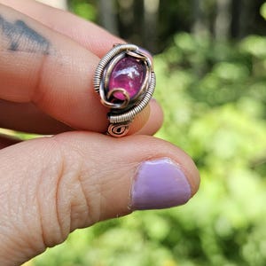 May include: A pink gemstone ring with a copper wire setting. The ring is on a finger with purple nail polish.