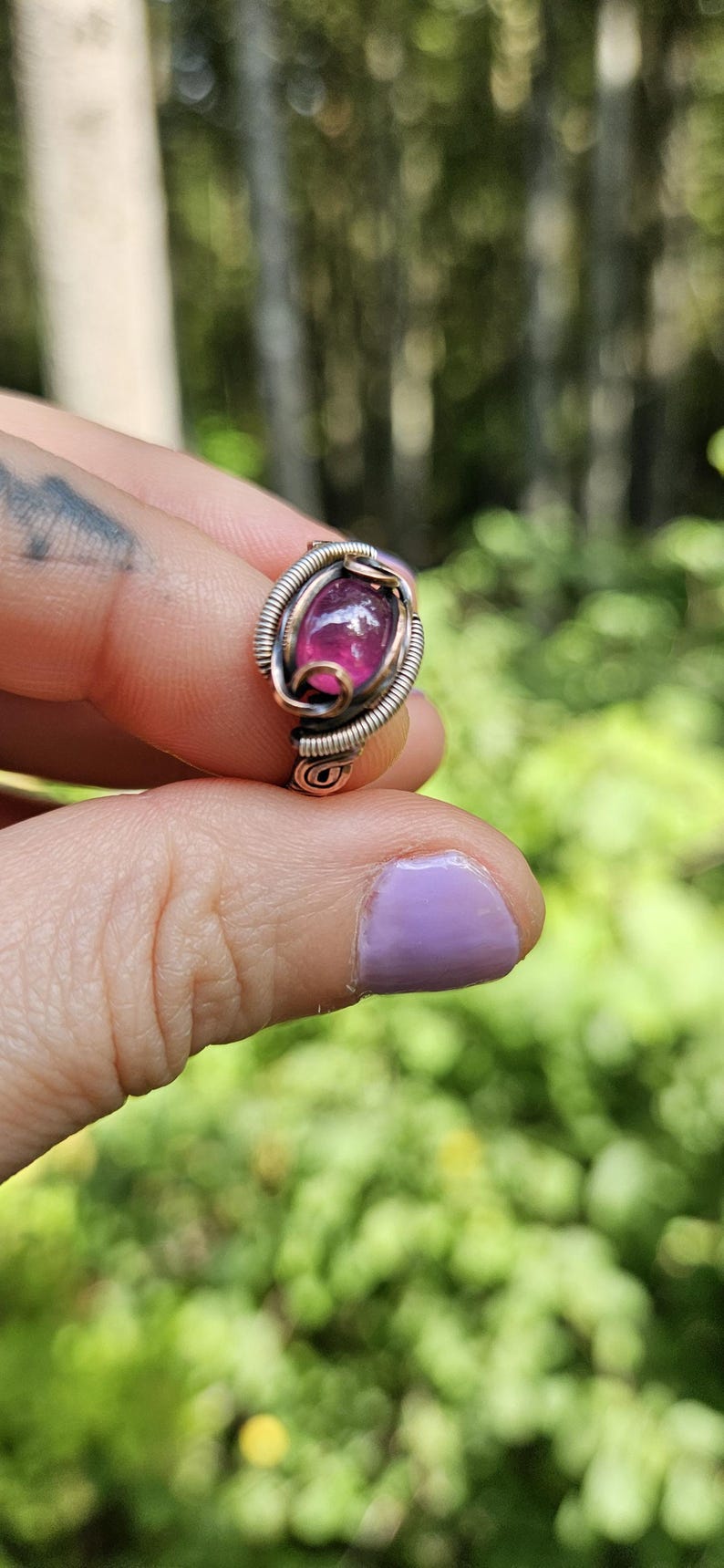 May include: A pink gemstone ring with a copper wire setting. The ring is on a finger with purple nail polish.