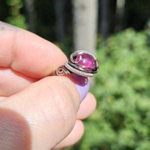 May include: A silver wire wrapped ring with a pink gemstone. The ring is on a finger and the background is blurred green foliage.