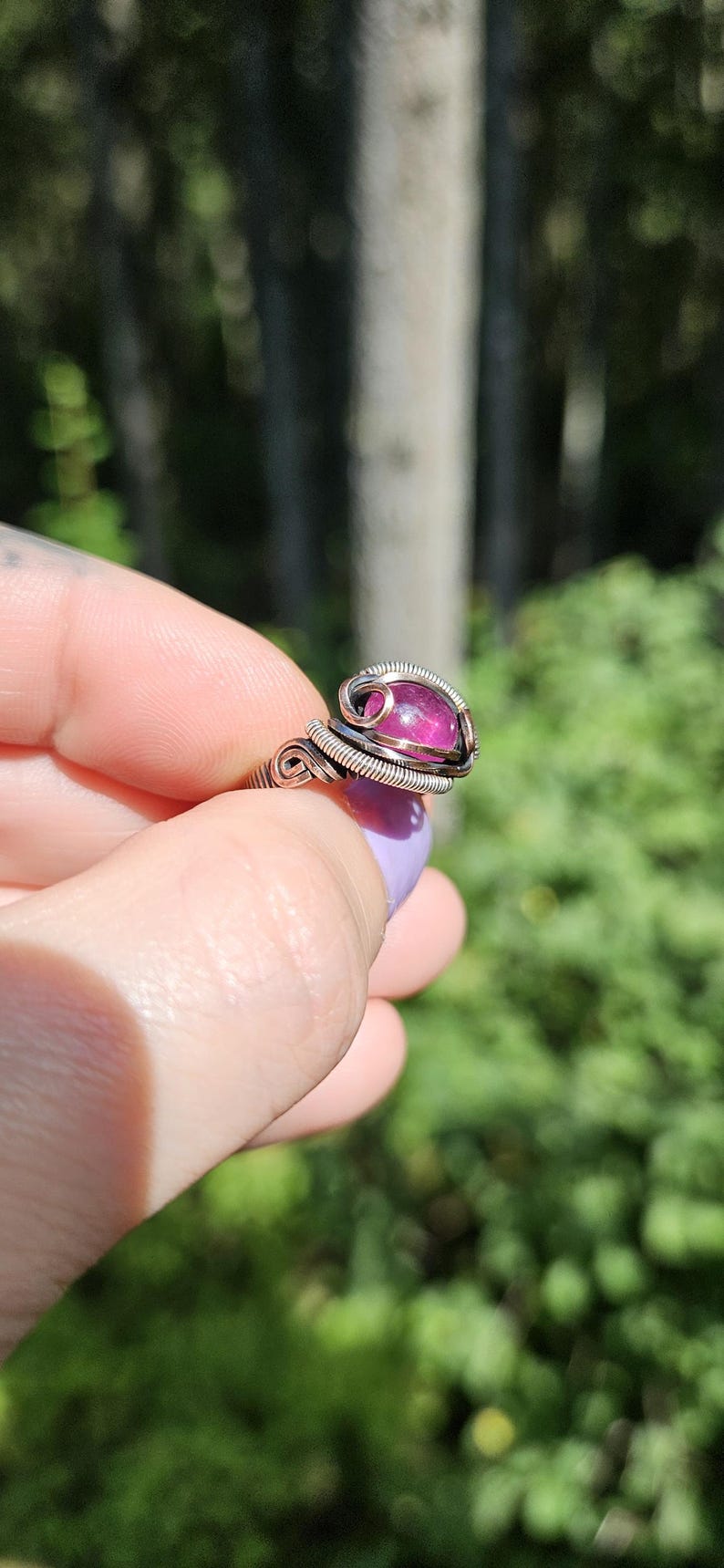 May include: A silver wire wrapped ring with a pink gemstone. The ring is on a finger and the background is blurred green foliage.