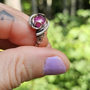 May include: A pink gemstone ring with a copper wire setting. The ring is on a finger with purple nail polish.