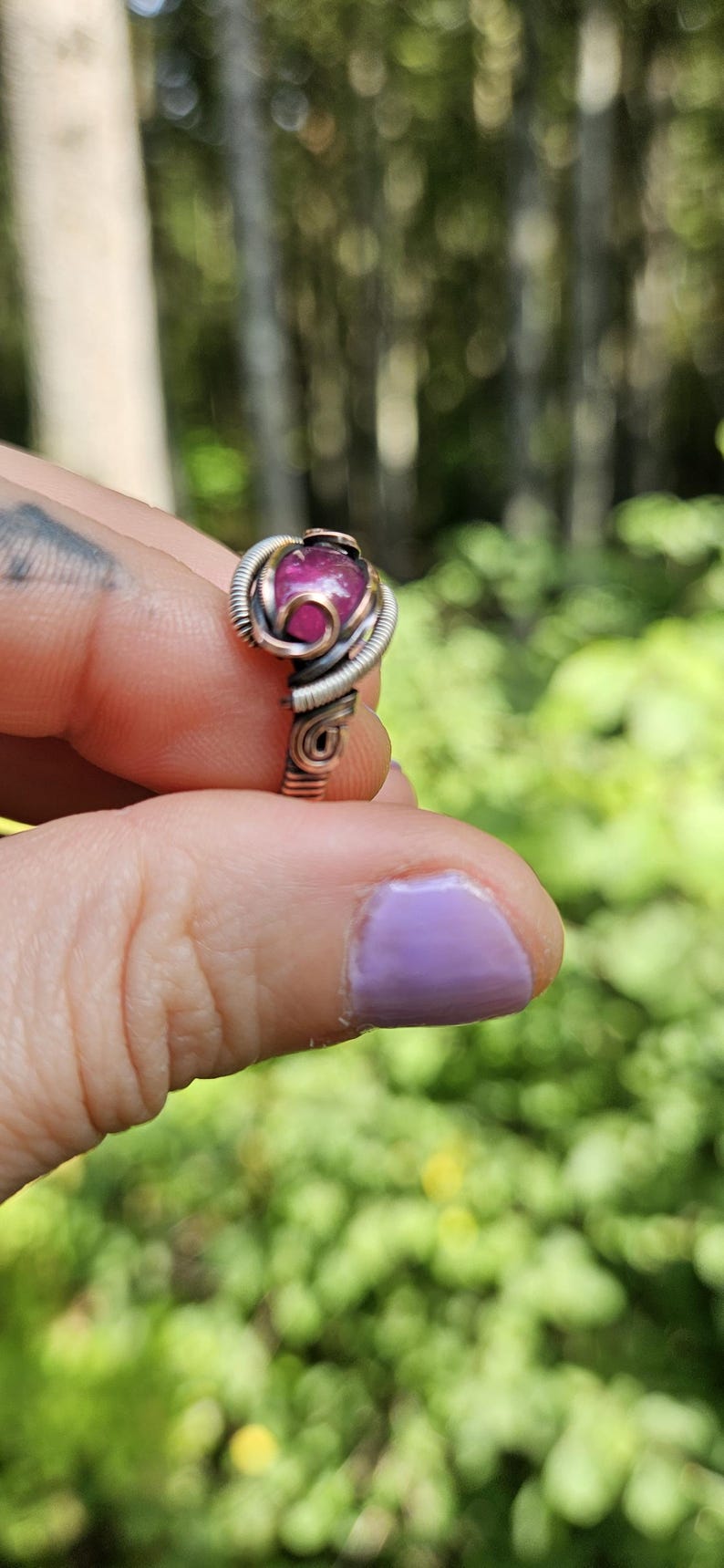 May include: A pink gemstone ring with a copper wire setting. The ring is on a finger with purple nail polish.