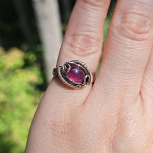 May include: A copper wire wrapped ring with a purple gemstone. The ring is on a person's finger.