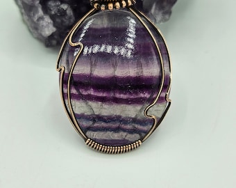 May include: A purple and white fluorite gemstone pendant wrapped in copper wire with a smaller blue gemstone bead at the top.