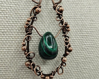 May include: A copper wire pendant with a green malachite stone. The pendant is made of copper wire that is twisted and shaped into a teardrop shape. The malachite stone is set in the center of the pendant and is surrounded by copper wire.