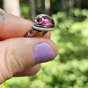 May include: A pink gemstone ring with a copper and silver wire wrap. The ring is set on a finger with purple nail polish.