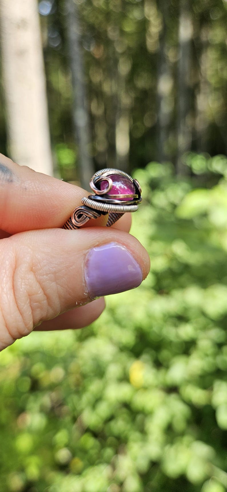 May include: A pink gemstone ring with a copper and silver wire wrap. The ring is set on a finger with purple nail polish.