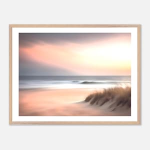 Puede incluir: Impresión enmarcada de un paisaje de playa al atardecer. La imagen tiene un efecto suave y borroso, con una paleta de colores pastel de rosa, naranja y gris. El océano se encuentra con la arena, con hierba de duna en primer plano.
