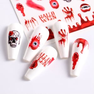 May include: White nail art with Halloween-themed designs. The designs include a skull, an eyeball, a bloody handprint, and dripping blood. The word "HALLOWEEN" is also featured on the nail art.