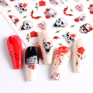 May include: A sheet of nail art stickers with skull and rose designs, along with several painted nail tips. The nail tips feature red, black, and nude colors, with skull, rose, and blood splatter designs.