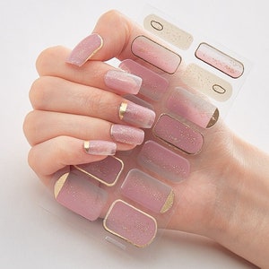 May include: A sheet of pink and white nail polish stickers with gold glitter accents. The stickers are designed to be applied to fingernails and feature a gradient design.