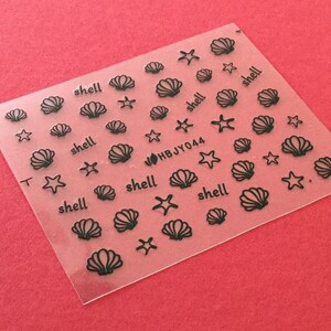 May include: A sheet of clear nail decals with black outlines of seashells and starfish. The sheet has the text "HBJY044" and "shell" repeated multiple times.