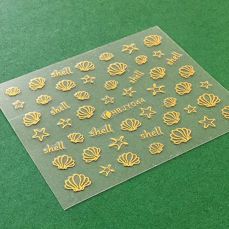 May include: A sheet of clear nail decals with gold seashells and stars. The sheet has the text "HBJY044" and "shell" repeated multiple times.