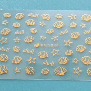 May include: A sheet of gold-colored nail stickers with images of seashells and starfish. The sheet has the text "shell" repeated multiple times and the code "HBJY044".