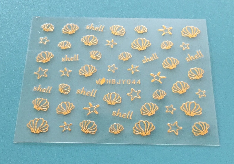 May include: A sheet of gold-colored nail stickers with images of seashells and starfish. The sheet has the text "shell" repeated multiple times and the code "HBJY044".