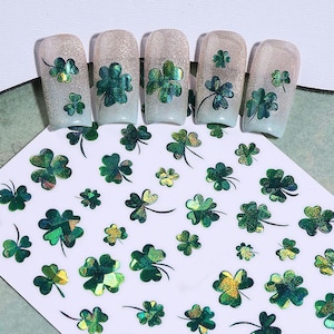 May include: Nail art stickers featuring green and gold shamrocks. The stickers are in various sizes and designs, with some applied to artificial nails. The nails have a glittery, iridescent finish. The shamrocks are a symbol of luck and are often associated with St. Patrick's Day.