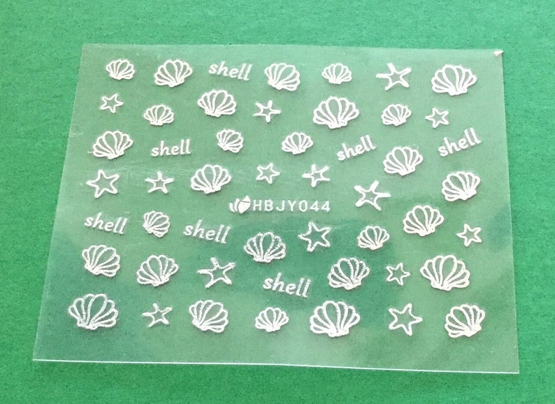 May include: A sheet of white nail decals with images of seashells and starfish. The sheet has the text "shell" repeated multiple times.