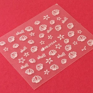 May include: A sheet of white nail decals with images of seashells and starfish. The sheet has the text "HBJY044" and the word "shell" repeated multiple times.